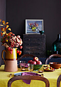 Dining room with dark purple walls and a autumnal table setting with golden yellow tablecloth and seasonal flowers and fruit