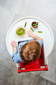A child in a high chair from above. Snacks on the high chair tray, mashed avocado with cucumber sticks and cut tomatoes