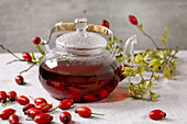 Rose hip berries herbal tea in glass teapot on white linen table cloth with wild autumn berries around