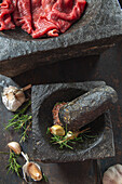 Rohes Rinderfilet auf schwarzer Steinplatte davor Mörser mit Kräutern und Knoblauch