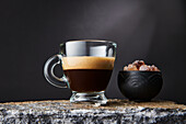 Transparent glass of strong black coffee with foam near bowl of brown sugar cubes on black background