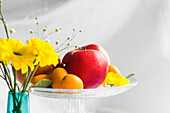 Äpfel, Mandarinen und Kumquats auf Glasständer, im Vordergrund gelbe Gerbera