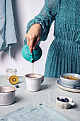 Crop anonymous female pouring brewed tea from teapot into mug for breakfast with granola