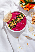 Beetroot hummus garnished with chickpea served on fabric background with bread and fresh carrot and cucumber sticks