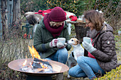 Two women with mulled wine and dog squatting around fire pit