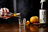 A Japanese sake with a cinammon stick, star anise and an orange peel being poured by a bartender