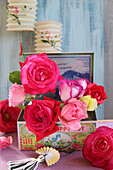 Kleine Glasvasen mit Rosen in Vintage Dose, darüber antike Laternen aus Papier