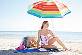 Junge blonde Frau im Badeanzug sitzt unter Sonnenschirm am Strand