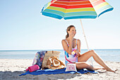 Junge blonde Frau im Badeanzug cremt sich ein unter Sonnenschirm am Strand