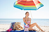 Young blonde woman in a bathing suit applying sunscreen under a parasol on the beach