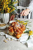 Colomba - traditional italian easter cake with almonds, woman sprinkling powdered sugar, easter table with mimosa in a vase