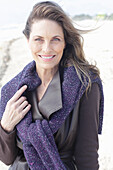 A long-haired woman wearing a dark coat with a jumper over her shoulders on the beach