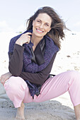 Long-haired woman in a dark coat with a sweater over her shoulders and barefoot on the beach