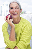 A grey-haired woman wearing a greenish-yellow knitted jumper holding an apple
