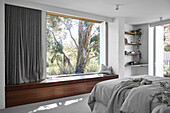 Doppelbett im Schlafzimmer mit weiß gestrichener Ziegelwand, Eingebauter Sitzbank aus Holz vor Panoramafenster