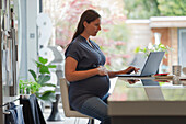 Pregnant woman working from home at laptop in kitchen