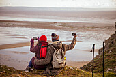 Hiker couple with phones on cliff overlooking winter beach