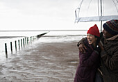 Happy couple hugging under umbrella on wet winter beach