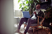 Mature man with tea using laptop in armchair at home