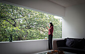 Young woman using smart phone at window with view of trees