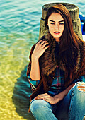 A young brunette woman wearing a cardigan with a fur collar and jeans at a lake
