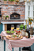 Gedeckter Tisch mit Pizza auf der Terrasse, im Hintergrund Holzofen