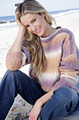 Young blond woman in knitted jumper with colour gradient and jeans by the sea