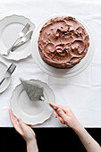 Whole chocolate cake covered in chocolate buttercream, on white cake stand, with hands holding a vintage cake slice.