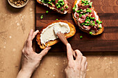 Hands spreading ricotta cheese on a crusty piece of bread next to pea, prosciutto, and ricotta crostinis