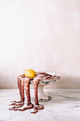 An octopus and a lemon in an old and decorated footed dish, the tentacles falling outside on a marble table