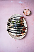 Bunch of fresh sardines placed on oval plate on pink table near bowl with salt