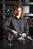 Focused female barkeeper in apron opening glass bottle of wine while standing at counter with wineglass during work in bar