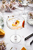 Elegant crystal glass with water decorated with flower petals and slices of orange in table with glassware and blooming branches