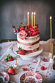 Delicious red velvet cake with burning candles decorated with fresh strawberries and flowers served on table during holiday celebration in kitchen
