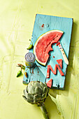 An artichoke and a slice of watermelon with the letter 'M' cut from it