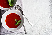 Top view of served spanish gazpacho tomato cream soup on light grey concrete background with fresh basil herb