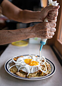 Hands of crop unrecognizable person pouring sauce on dish with fried eggs and nachos placed on counter in light kitchen