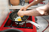High angle of crop anonymous person breaking raw eggs on hot frying pan on gas stove while preparing meal for breakfast in kitchen