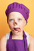 Astonished messy boy in purple chef uniform with sweet chocolate on mouth looking at camera sticking tongue out while standing on yellow background in studio