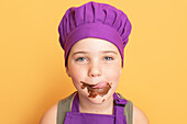 Cute dirty boy in chef uniform looking at camera while licking sweet chocolate off face against yellow background in studio