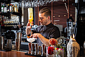 Concentrated young male bartender with trendy hairstyle in uniform pouring orange liquid into jigger while standing at counter and preparing alcohol cocktail with ice in bar