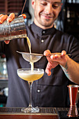 Crop bearded young male bartender in uniform pouring mixed cocktail from shaker into glass through strainer while standing at counter in modern bar