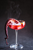 Alcoholic cocktail with ice cubes served with octopus tentacle and lemon slice placed on table against gray background in studio