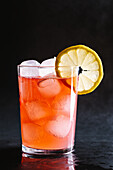Ice cubes pouring into glass with alcoholic red cocktail with lemon slice served on gray table against dark background in studio