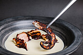 Tasty fried octopus tentacles on tongs served in bowl with white sauce on table against gray background in light kitchen