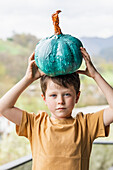 Adorable boy with teal pumpkin above head looking at camera while standing in rural area with mountains on blurred background