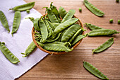 Wicker bowl full of ripe snow peas placed near napkin on wooden table at home