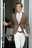 A young businessman wearing a brown jacket and light-coloured trousers stepping out onto the street