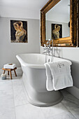 Free standing bathtub, gold framed mirror and painting above in light grey bathroom