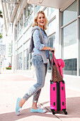Junge blonde Frau in Freizeitbekleidung mit pinkem Trolley und Hut